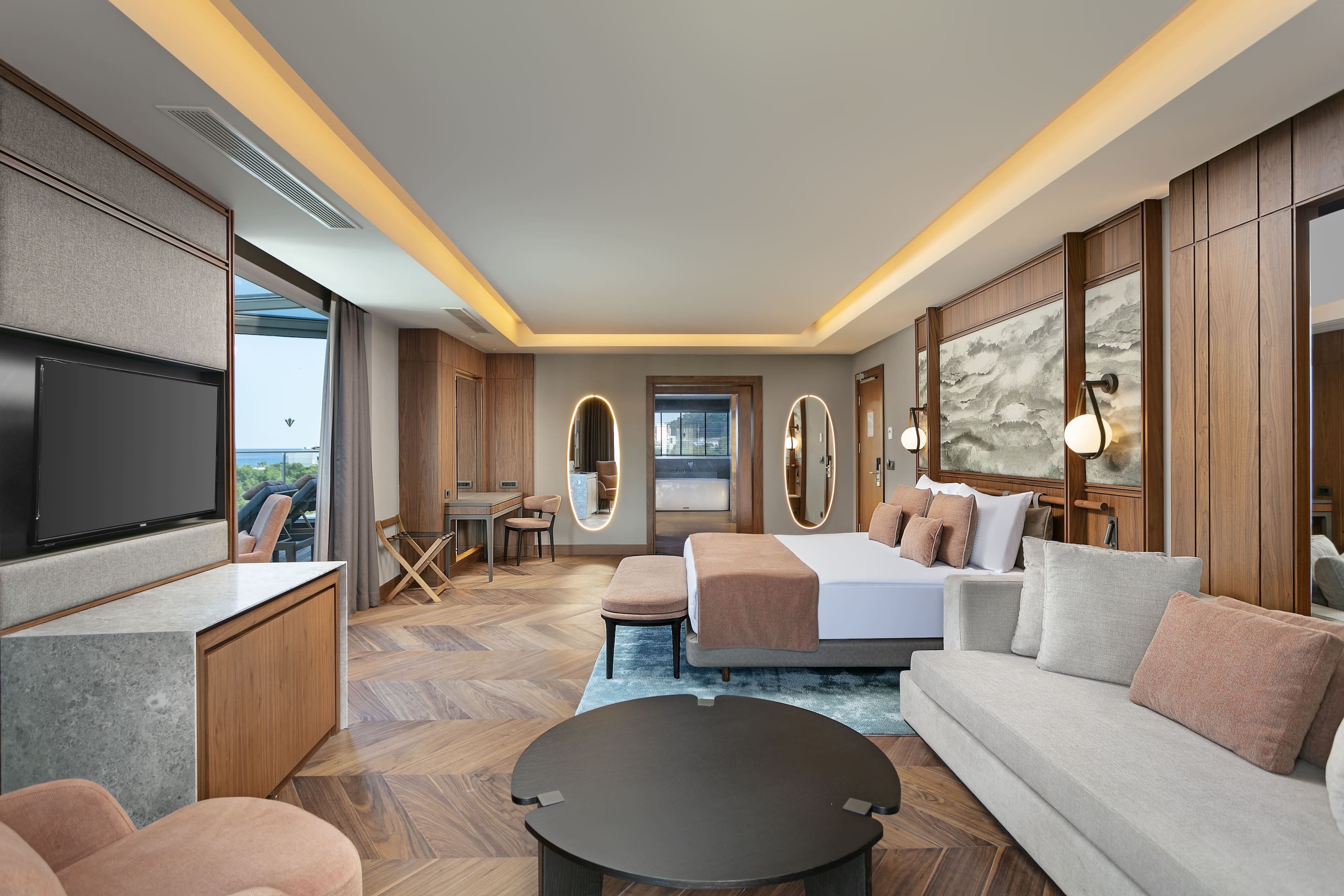 Weitläufiger Blick auf das moderne Hotelzimmer mit einem Kingsize-Bett, einem hellgrauen Sofa, einem runden Couchtisch, einem TV-Möbel und Blick auf den hellen Badbereich.