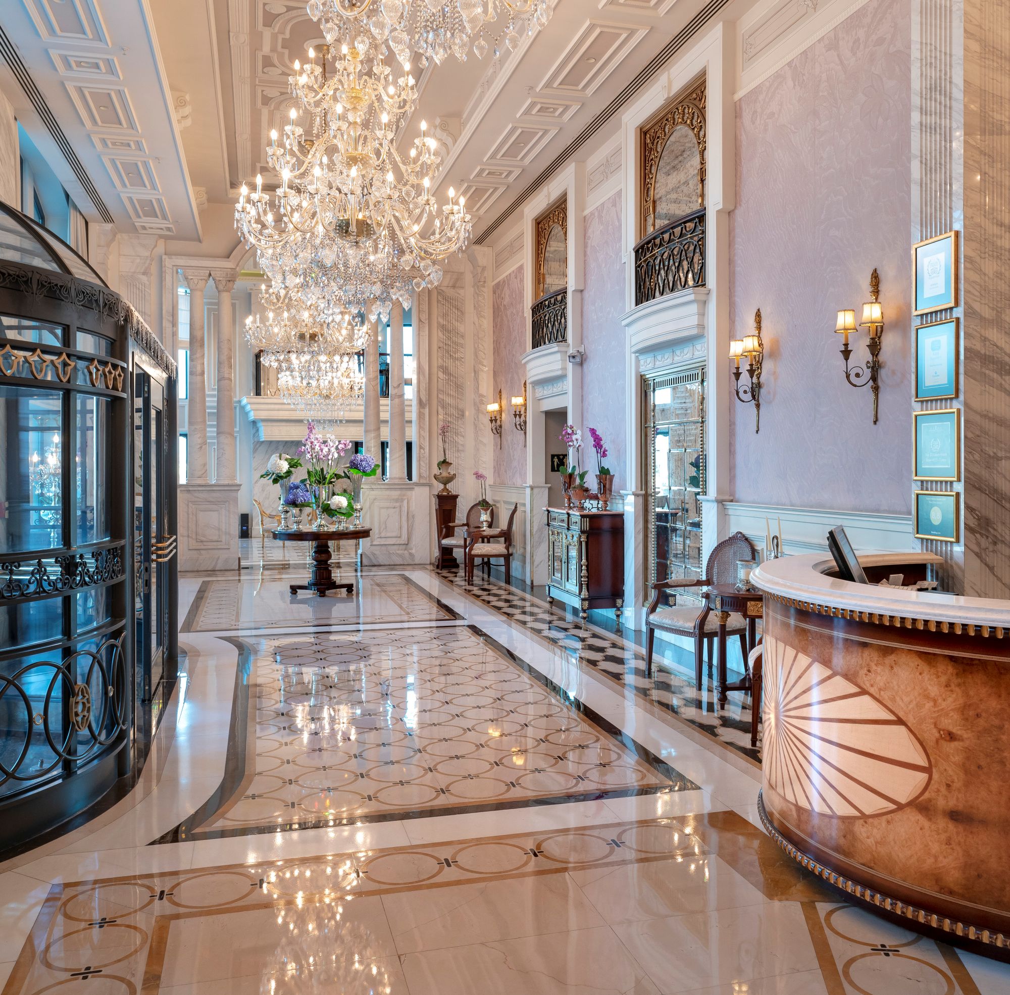 O luxuoso átrio do hotel Rixos Pera Istanbul apresenta tetos altos, lustres elegantes, pisos em mármore, um balcão de receção redondo e grandes janelas que permitem a entrada de luz natural.