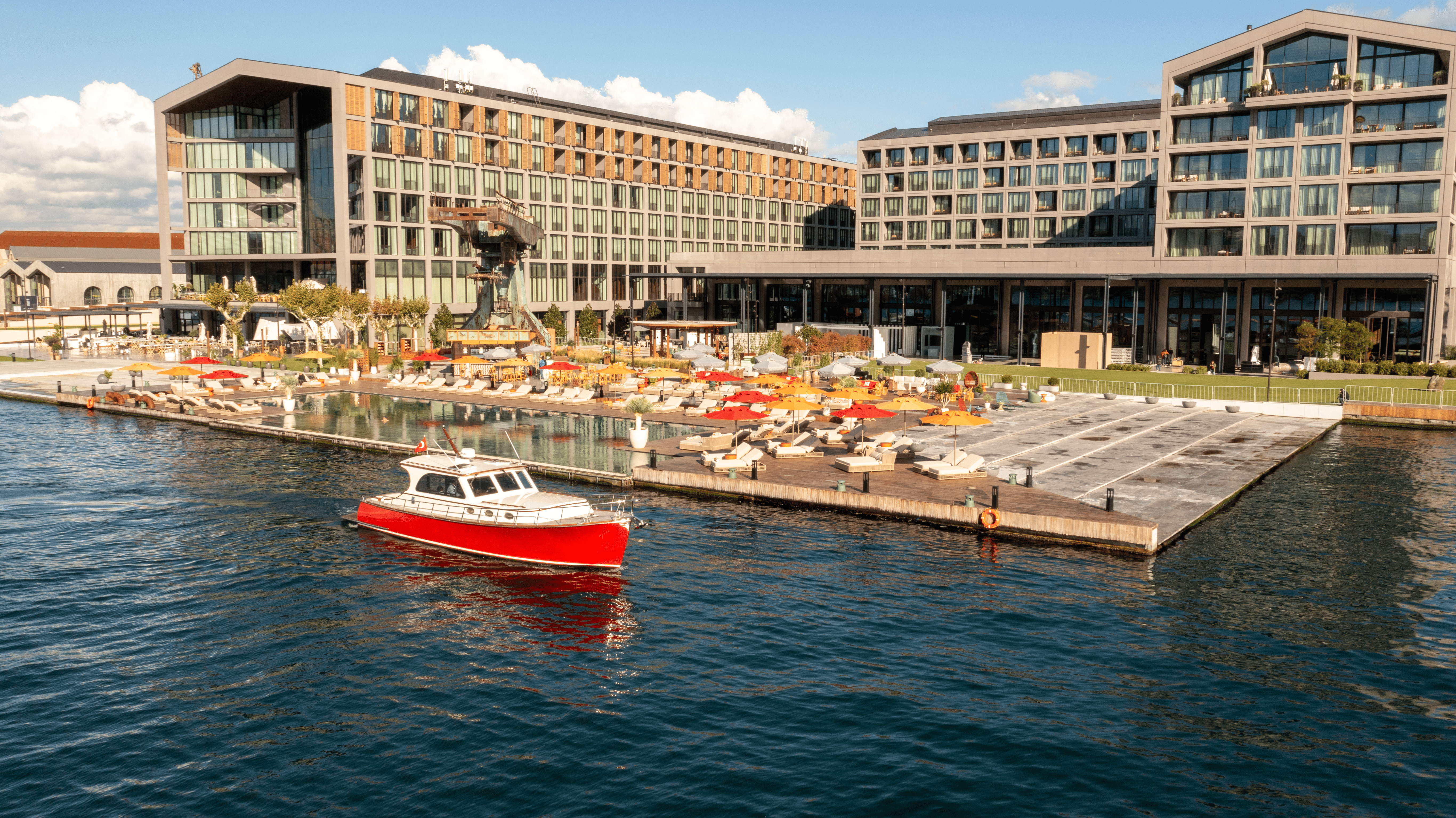 O imagine largă a complexului hotelier Rixos Tersane Istanbul, cu două clădiri moderne mari, cu vedere la o terasă pe malul apei, cu șezlonguri și umbrele de soare roșii și portocalii. O mică barcă roșie și albă navighează pe apa albastră din prim-plan.