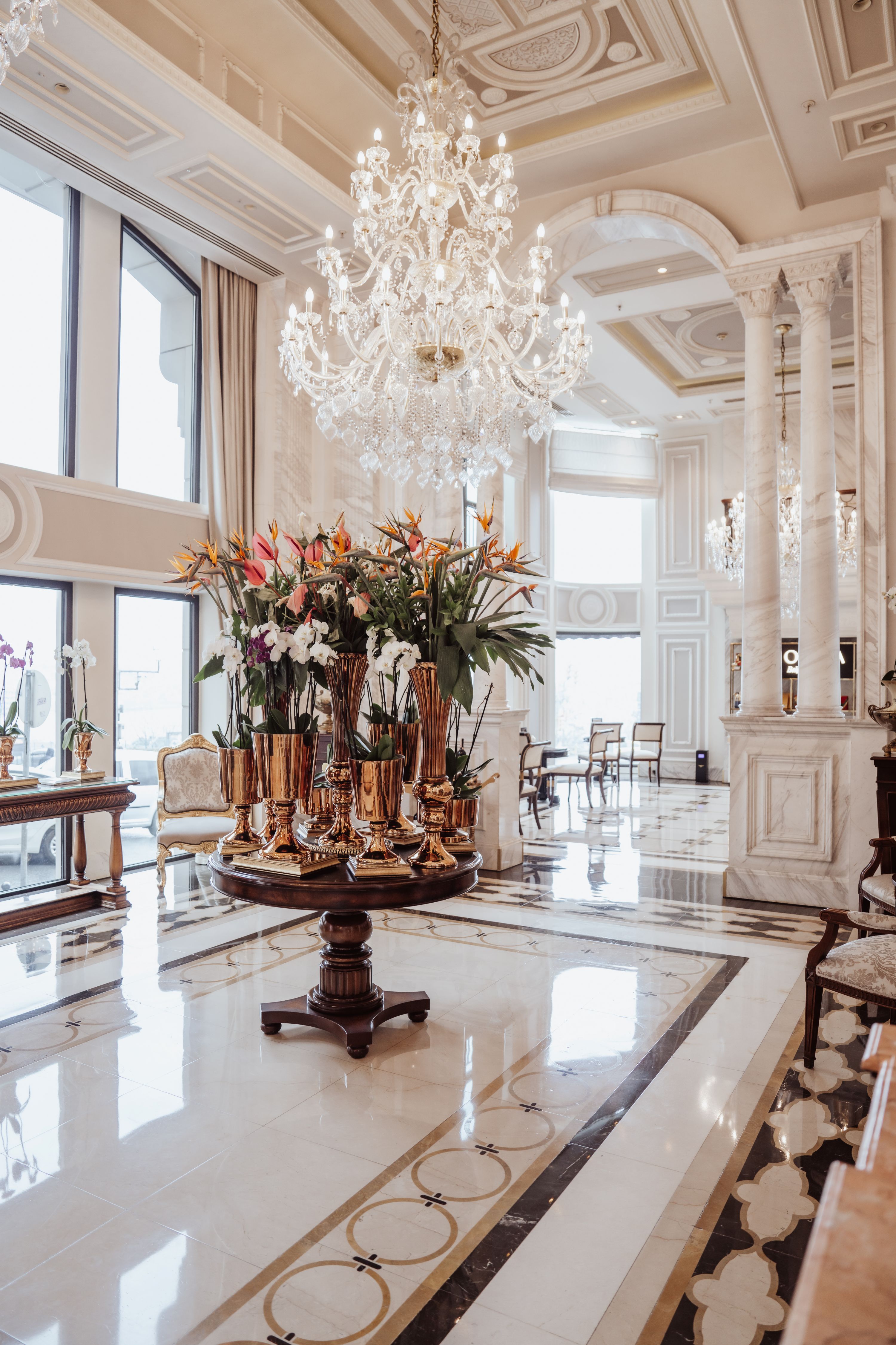 Роскошное, светлое лобби отеля Rixos Pera Istanbul с высокими потолками, высокими окнами, большой люстрой и центральным круглым столом, украшенным высокой цветочной композицией. В декоре использованы элегантные белые и золотые акценты