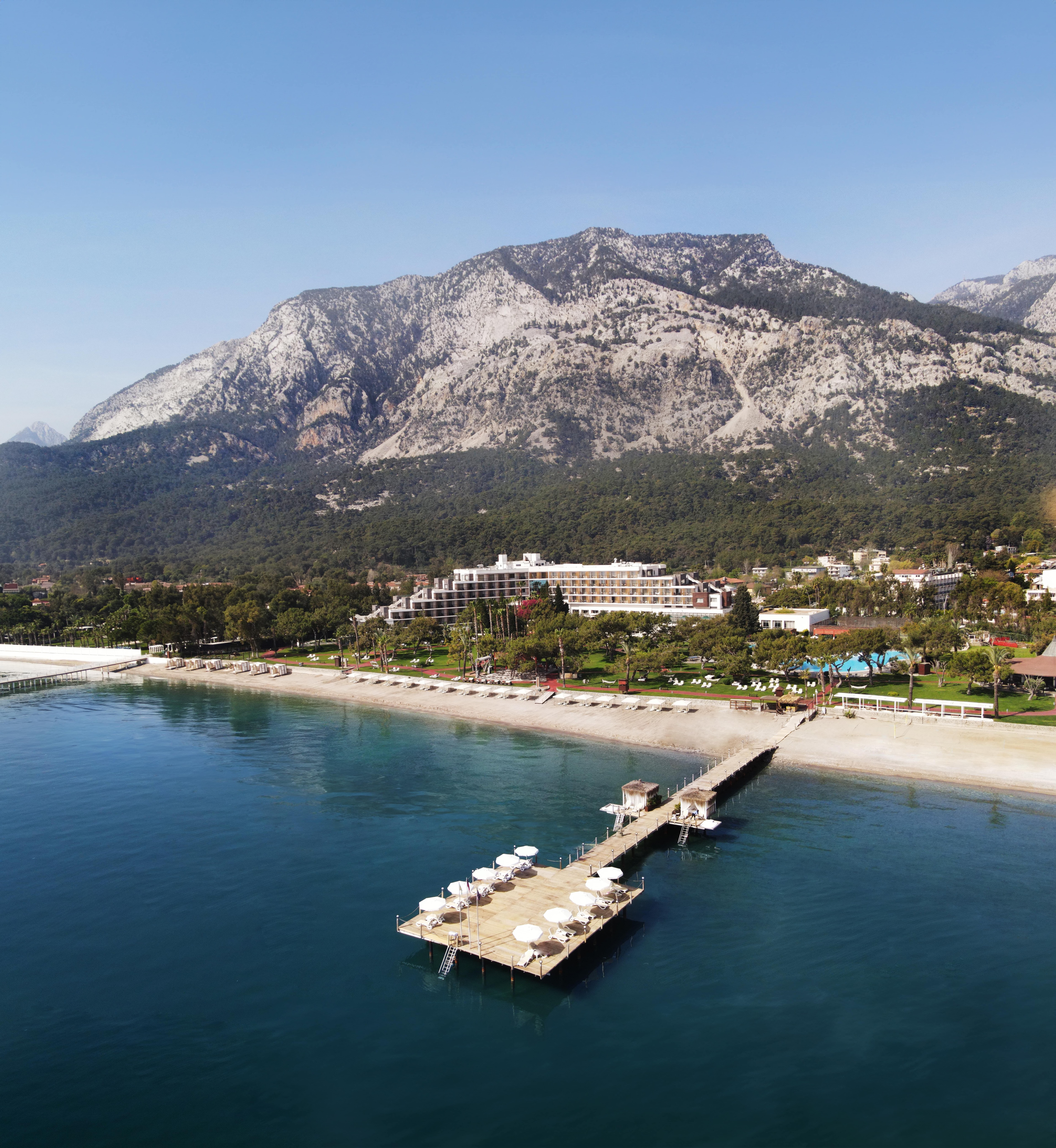 A wooden pier with white umbrellas extends over calm blue water toward a sandy beach, backed by the Rixos Beldibi hotel, green trees, and tall, rocky mountains under a clear sky