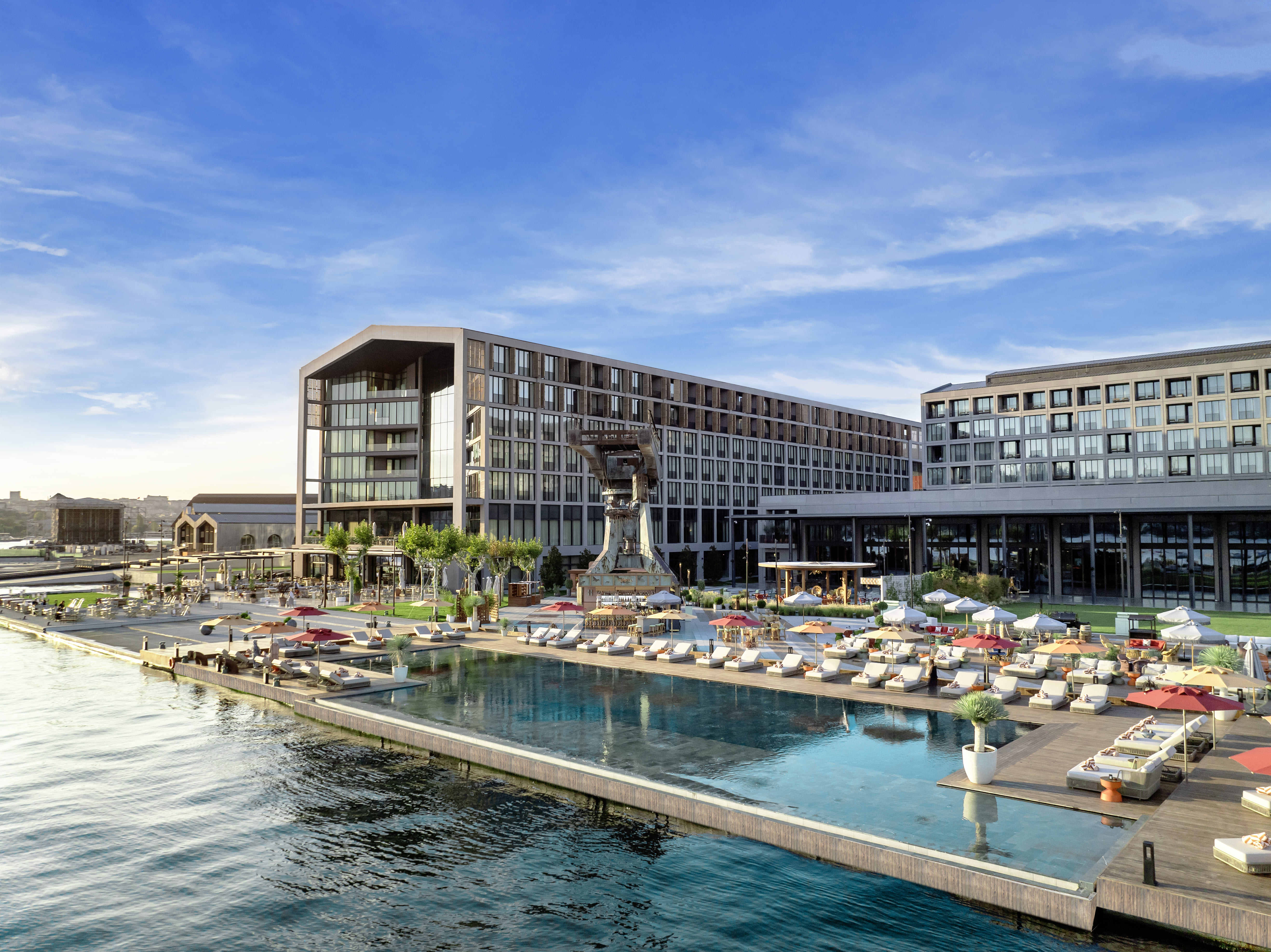 A modern waterfront hotel, Rixos Tersane Istanbul features large glass windows, an outdoor infinity pool, sun loungers, umbrellas, and landscaped gardens overlooking calm water under a clear blue sky.