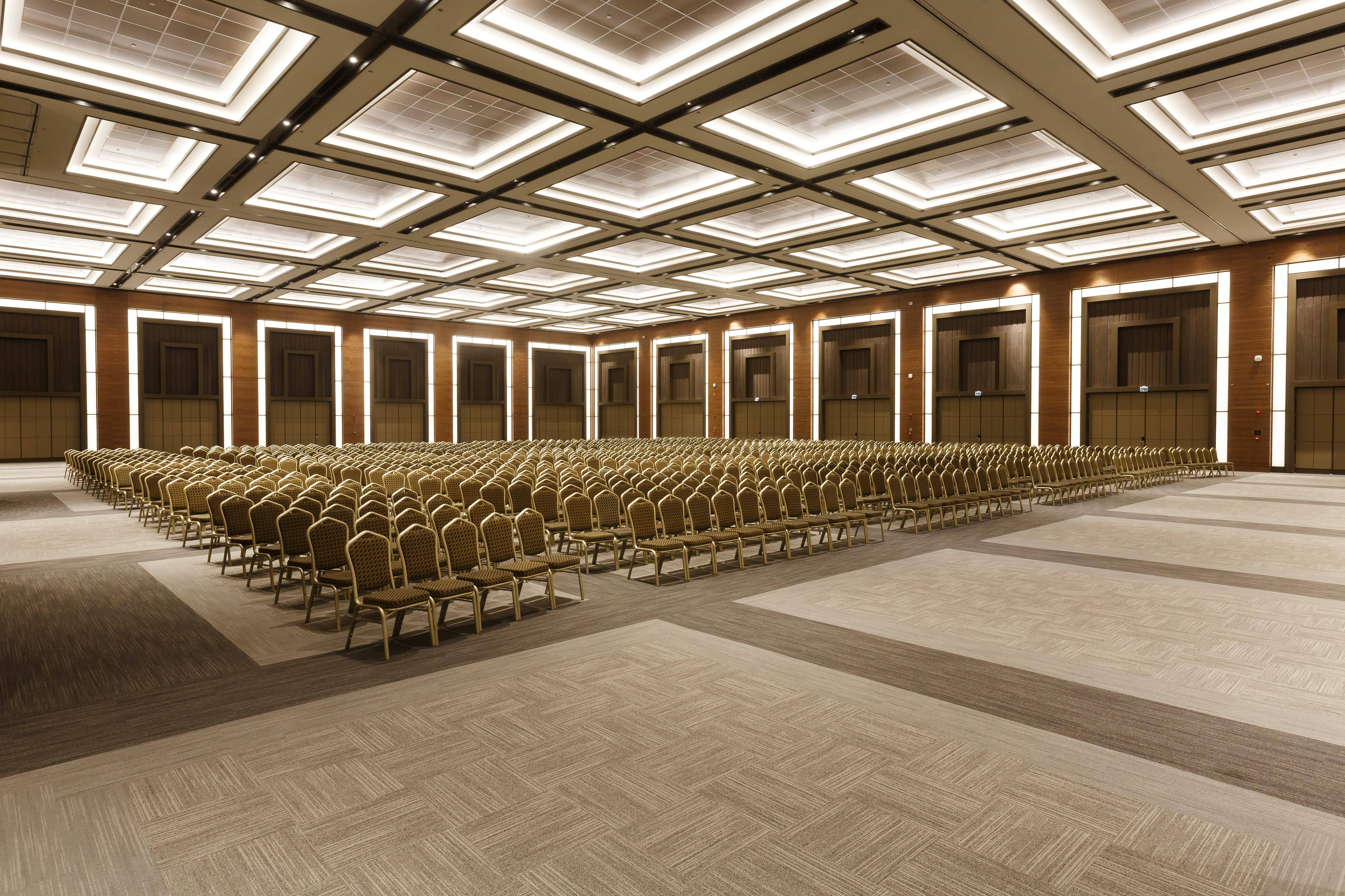 A wide, slightly low-angle view of the spacious Maximus Ballroom at Rixos Sungate, set up in a theater-style configuration for a large congress or meeting. Hundreds of gold and beige chairs are arranged in straight rows across the center of the hall. The room features a large, flat ceiling with many square, illuminated light panels. The walls are paneled in dark wood with evenly spaced vertical light strips, giving the Rixos venue a grand, modern look.