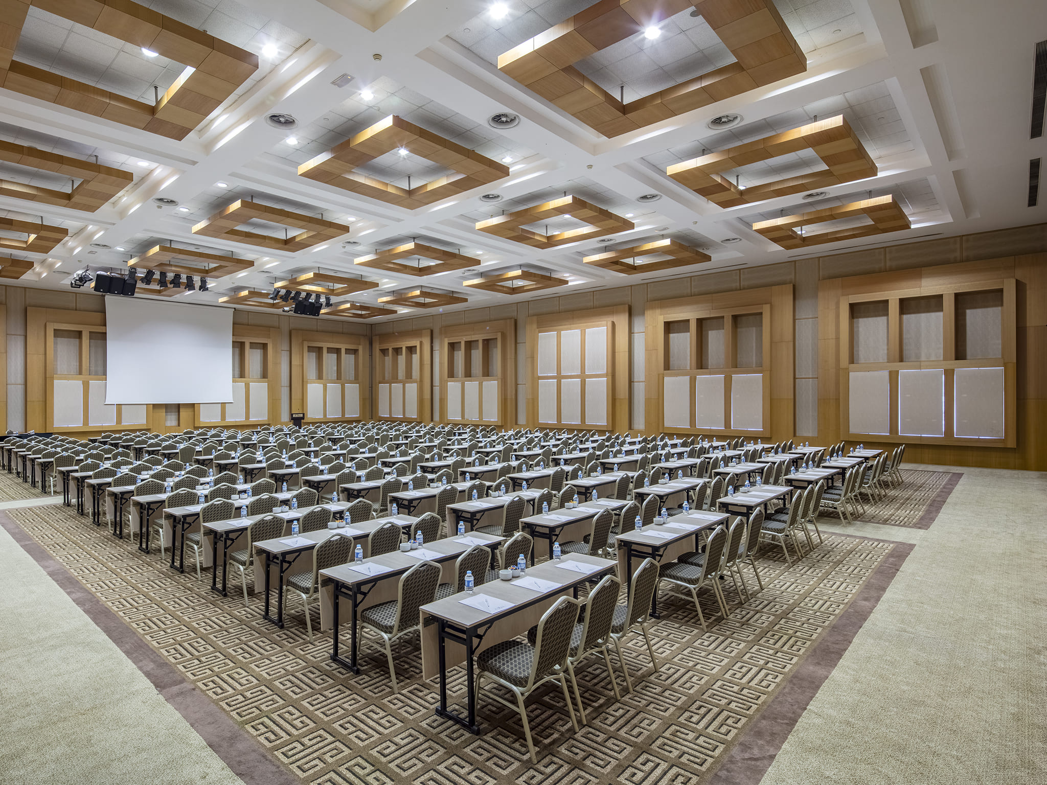A wide-angle image of the brightly lit Atlas Ballroom at Rixos Sungate, configured in a classroom setup for a large-scale congress or conference. Numerous rows of long tables, each set with water bottles and stationery, face a large projection screen and stage area. The room features light wood paneling on the walls and a contemporary ceiling design with suspended wooden frames and recessed lighting. The carpet has a bold, geometric pattern, defining the event space in this Rixos venue.