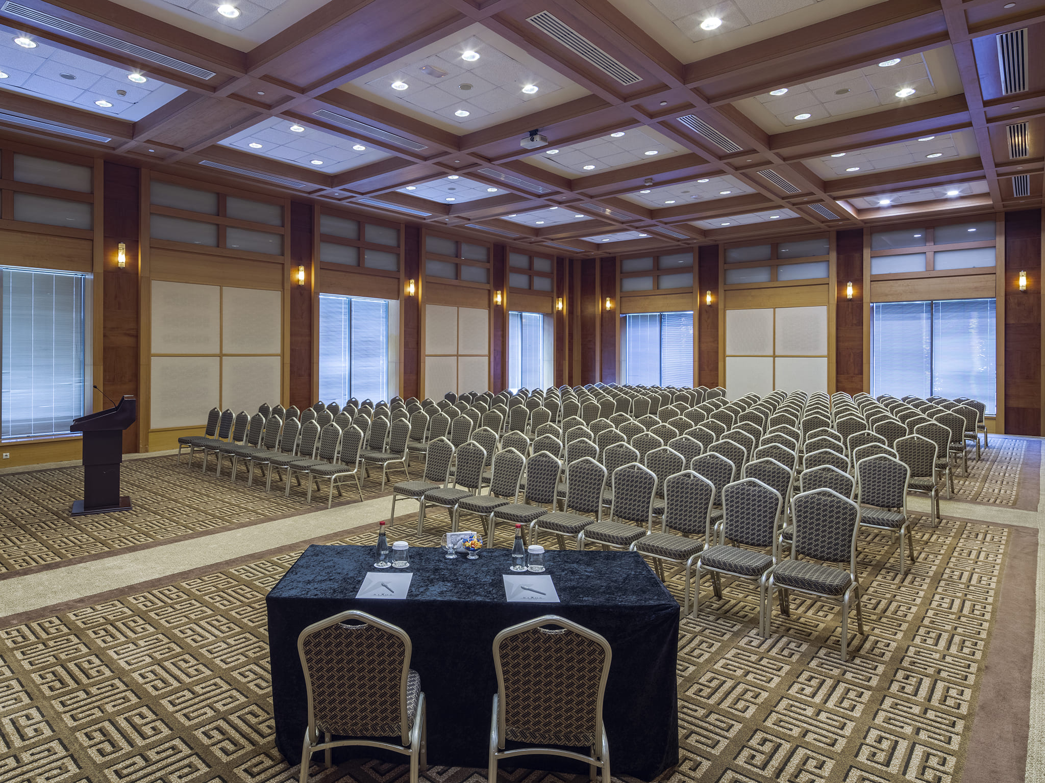 A slightly low-angle view of the Atria Meeting Room at Rixos Sungate, arranged in a theater-style setup for a congress or large presentation. Numerous rows of gray and brown patterned chairs face a dark wooden podium on the left. In the foreground, there is a black draped table with two chairs. The room is decorated with warm wooden ceiling panels and wall frames, interspersed with windows covered by blinds. The floor has a distinct brown and gold geometric carpet, characteristic of the Rixos conference fac