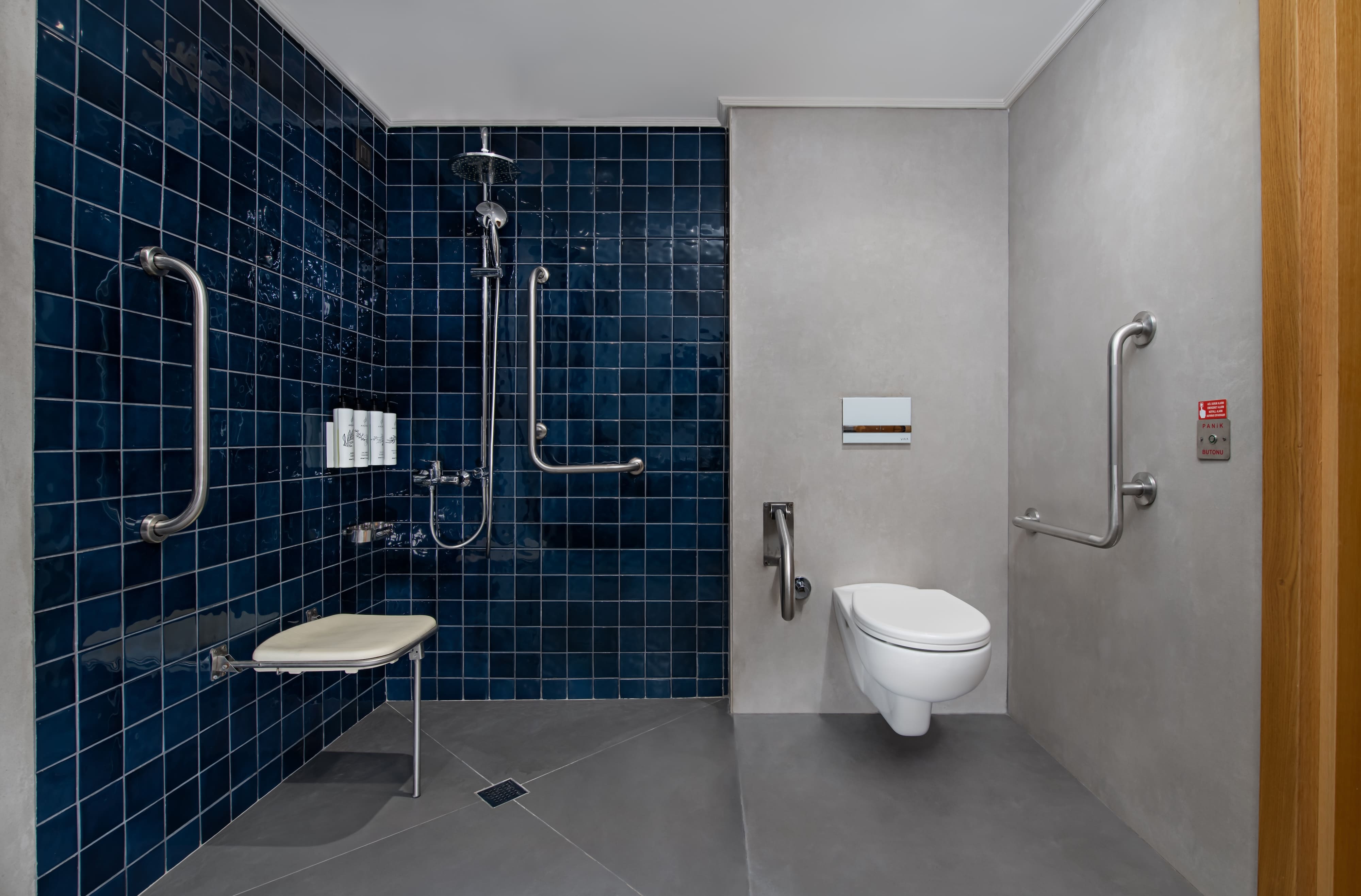 Accessible bathroom featuring a roll-in shower area with dark blue square tiles on the back wall and gray non-slip flooring. It includes a wall-mounted, folding shower seat, multiple grab bars for support, and an accessible wall-mounted toilet. An emergency pull cord is visible near the toilet