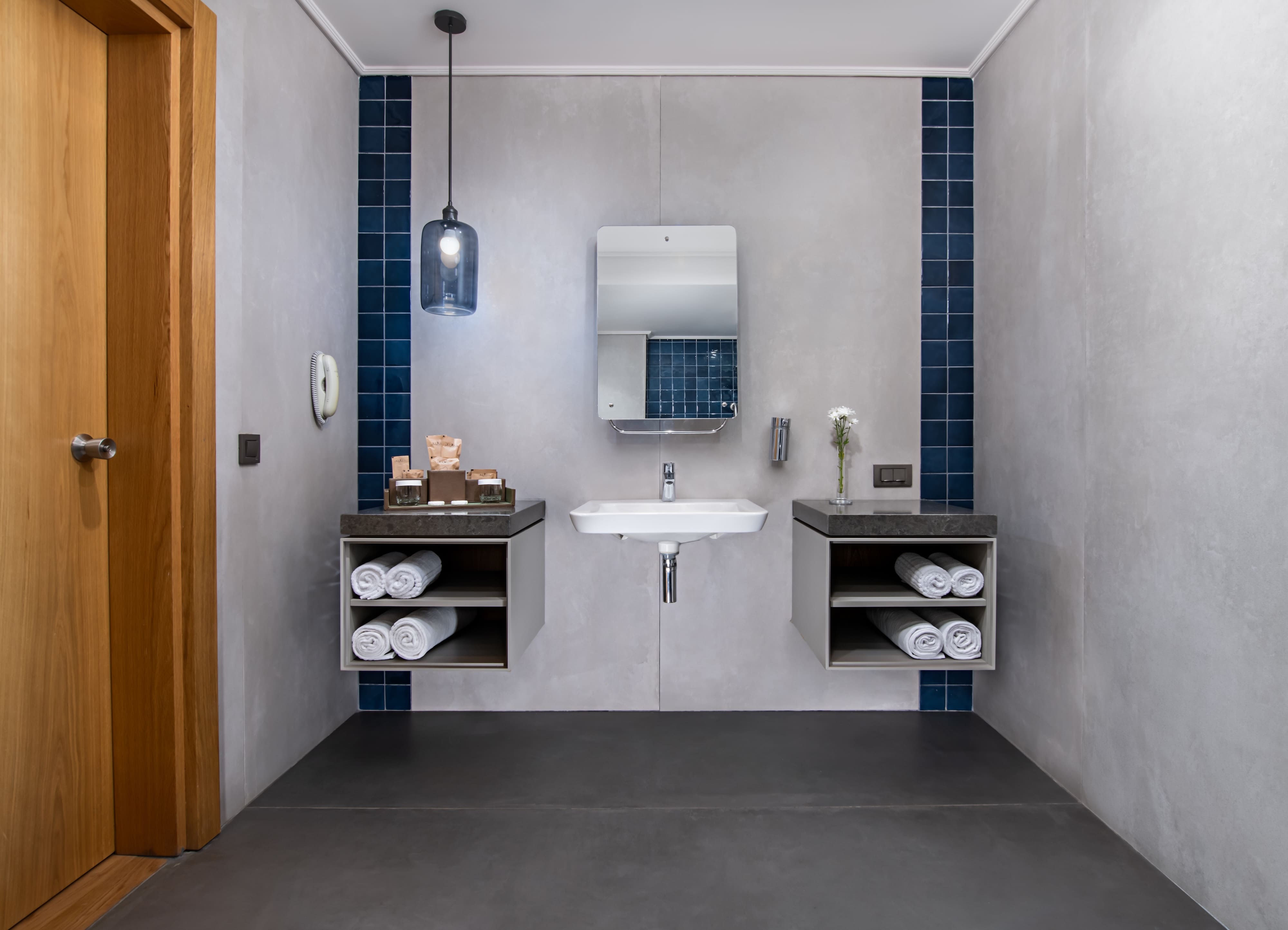 Accessible vanity area of the bathroom with a low, wall-mounted sink and a large, frameless mirror. The room has light gray walls and dark flooring. Storage is provided by two open shelving units beneath the counter, which hold rolled white towels. A light wooden door is visible to the left