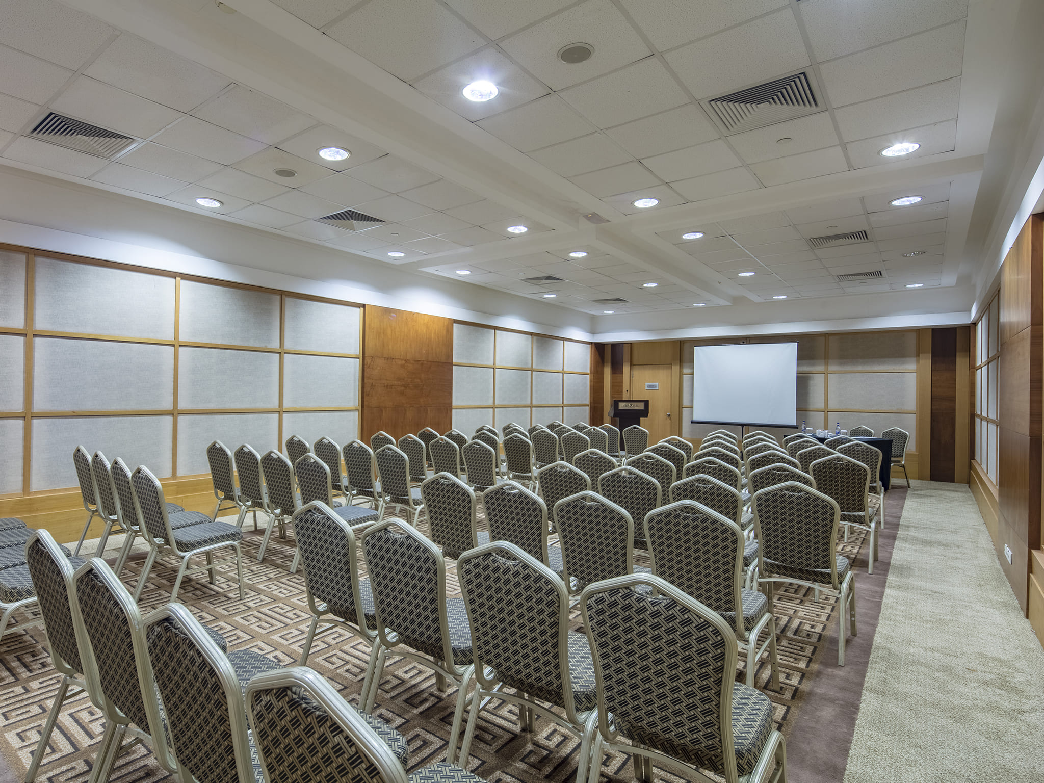 A photograph of the Maia Meeting Room at Rixos Sungate, configured in a hollow-square setup for a business meeting or small-scale congress. A long arrangement of tables is surrounded by gray, patterned chairs. The tables are set with water glasses, bottles, and stationery. A wooden podium and a projection screen are visible at one end of the room. The space features recessed lighting, beige walls, and a carpet with a geometric pattern, representing a professional setting at the Rixos hotel.