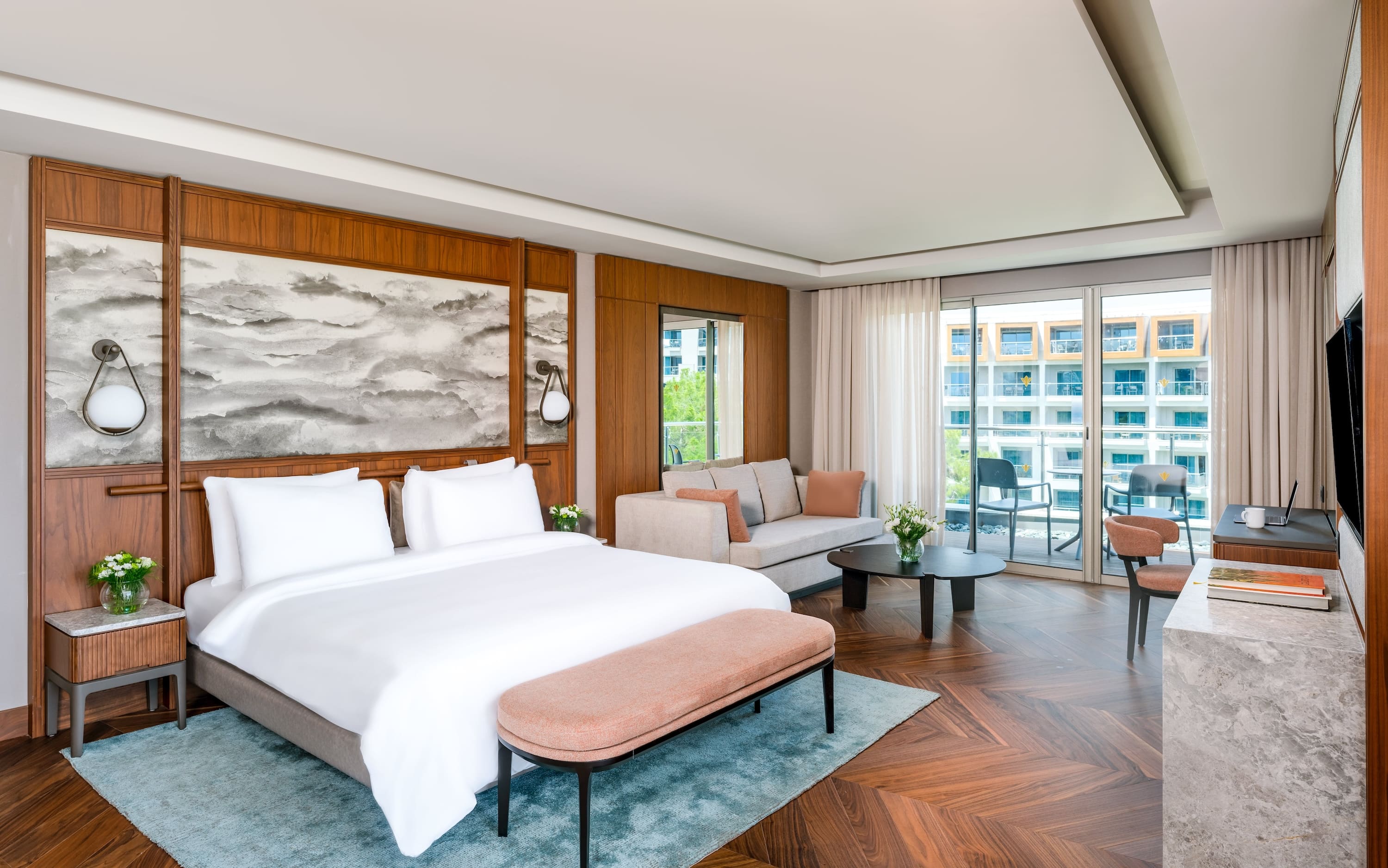 Modern hotel room with a large bed, light wood paneling, a small sofa area, and a glass door leading to a balcony.
