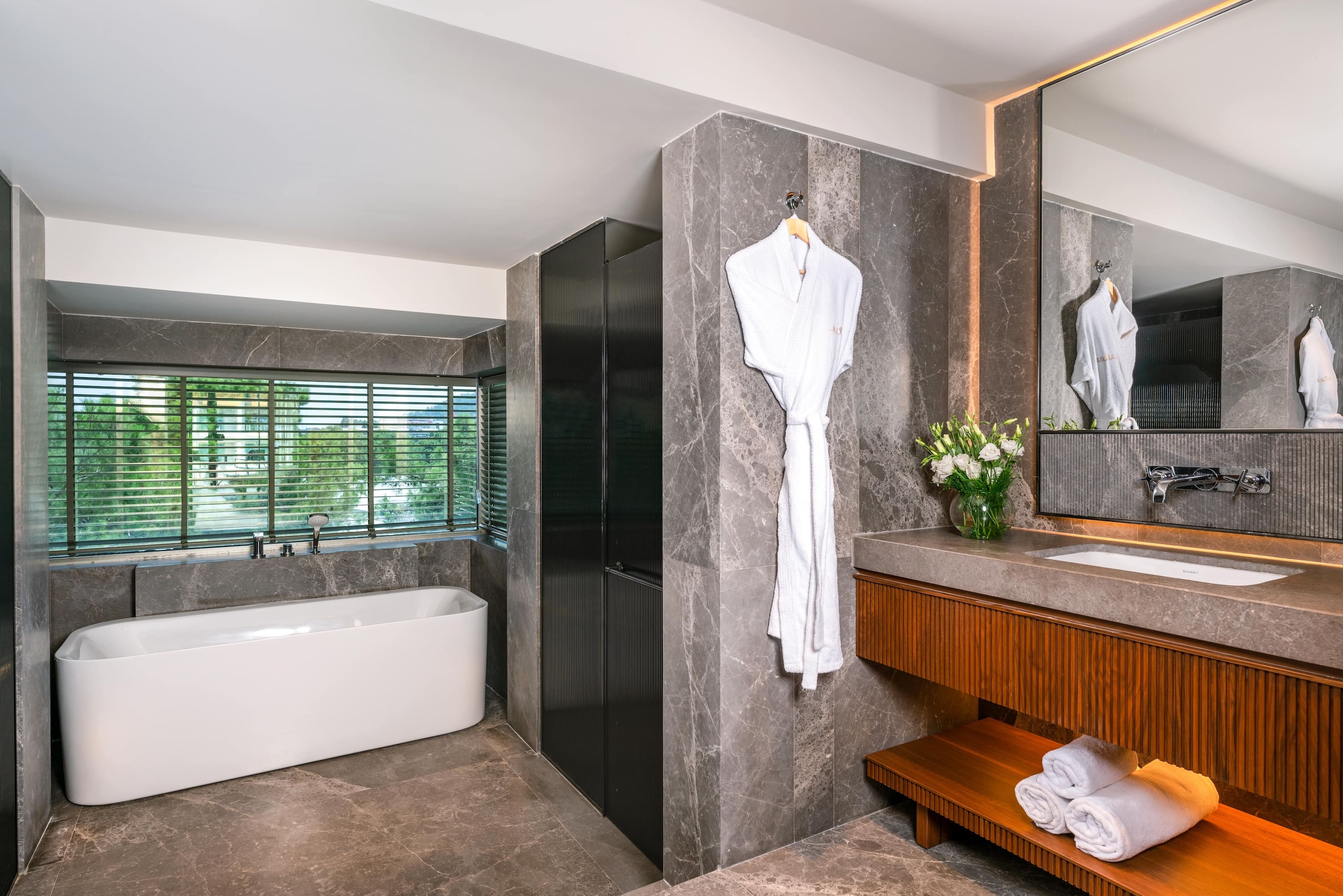 Spacious bathroom with dark grey marble walls, a freestanding white bathtub in front of a window, and a wooden vanity with a sink and fresh towels.