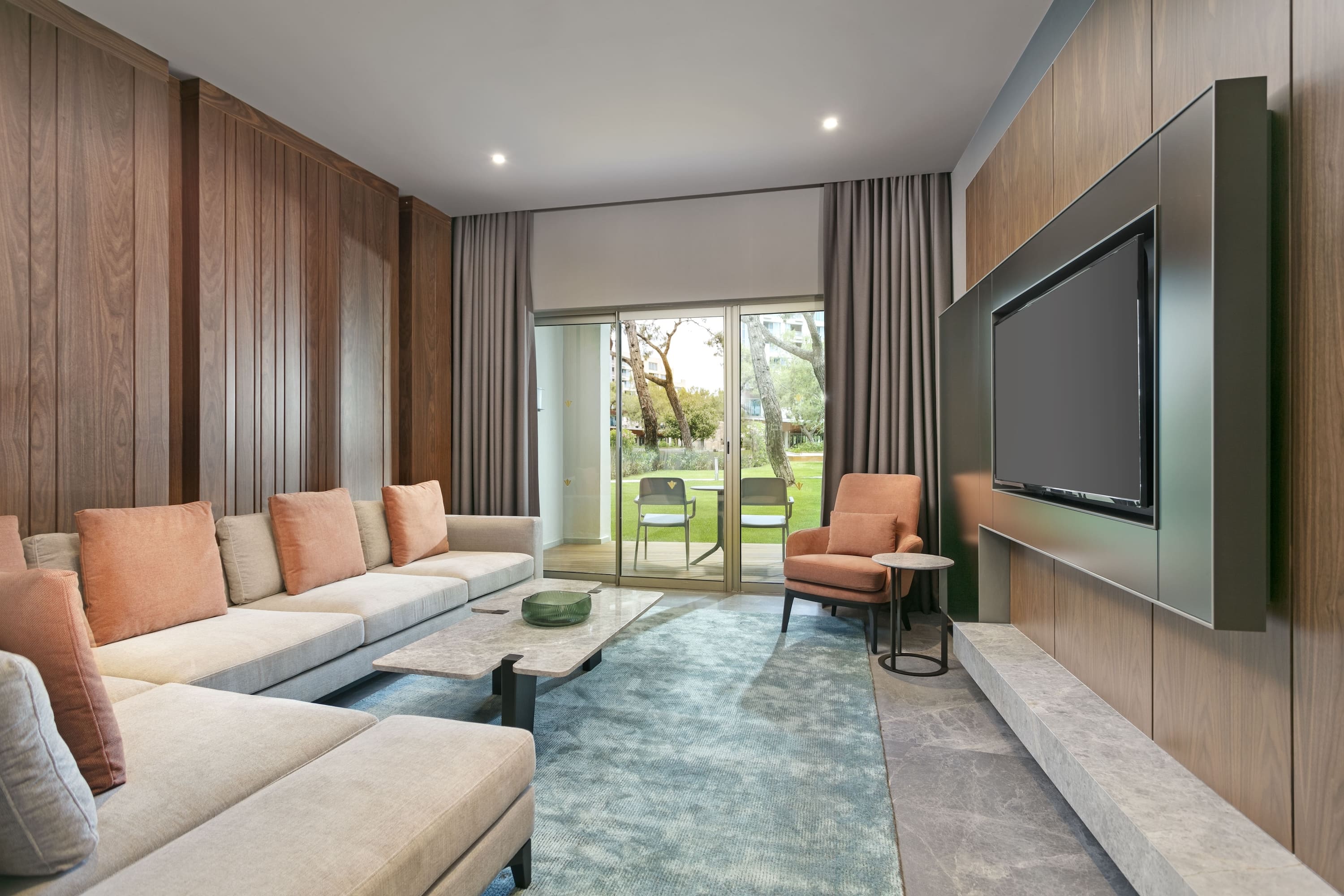 Living room area of the Marine Suite at Rixos Sungate, featuring a large L-shaped sofa with terracotta cushions, modern wood paneling, and a wall-mounted TV. Sliding glass doors provide natural light and direct access to a garden terrace