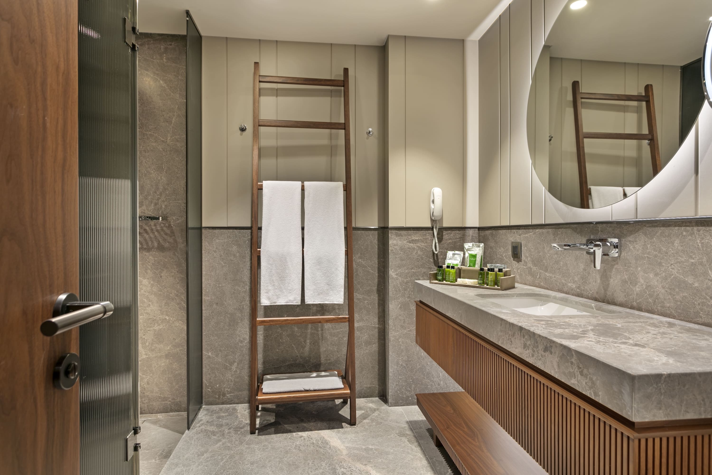 Luxury bathroom in the Marine Suite featuring grey marble walls and vanity, a wooden ladder towel rack, and a large round backlit mirror.