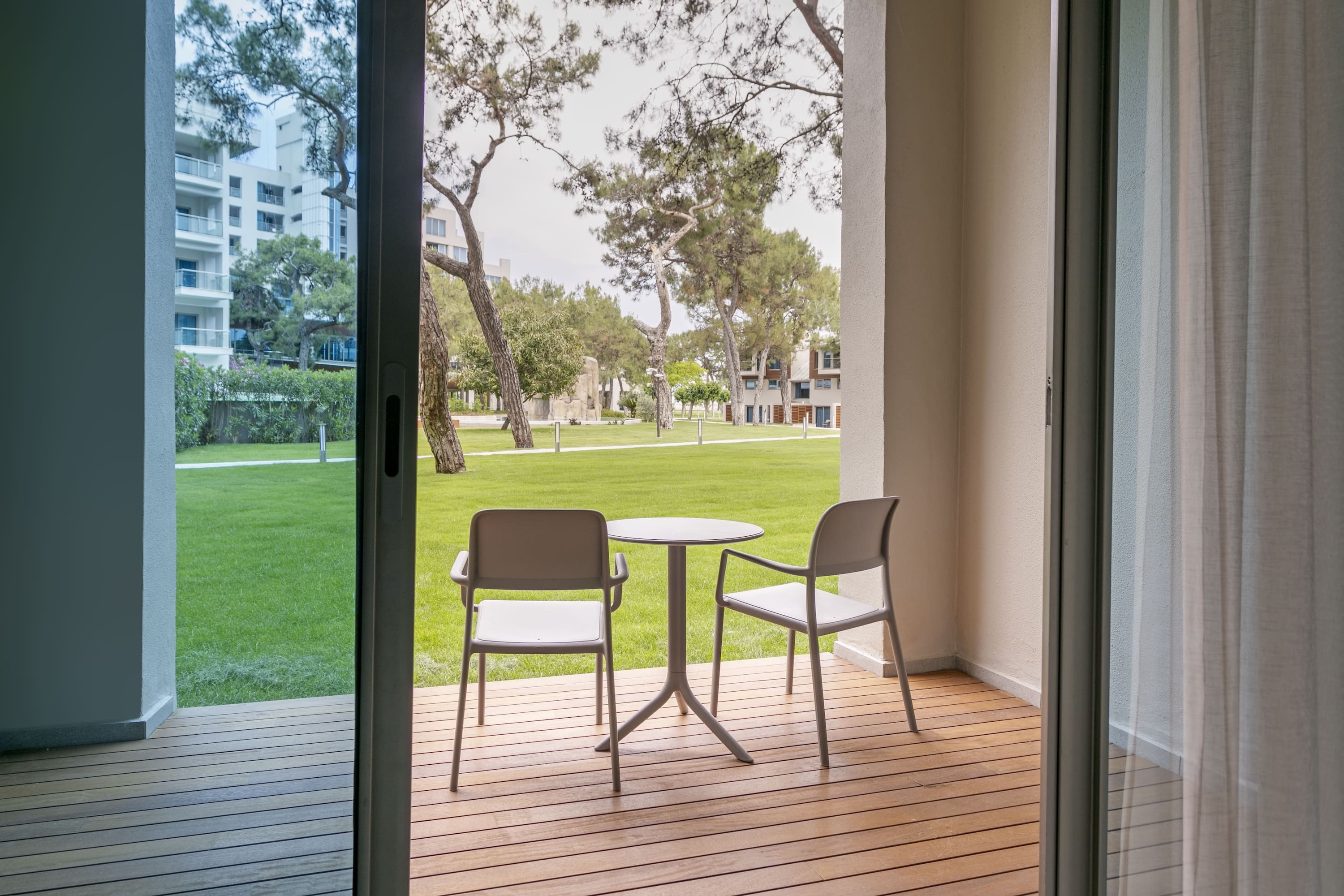 View from the Marine Suite terrace looking out through sliding glass doors onto a wooden deck furnished with a table and chairs, overlooking a lush green lawn and pine trees.