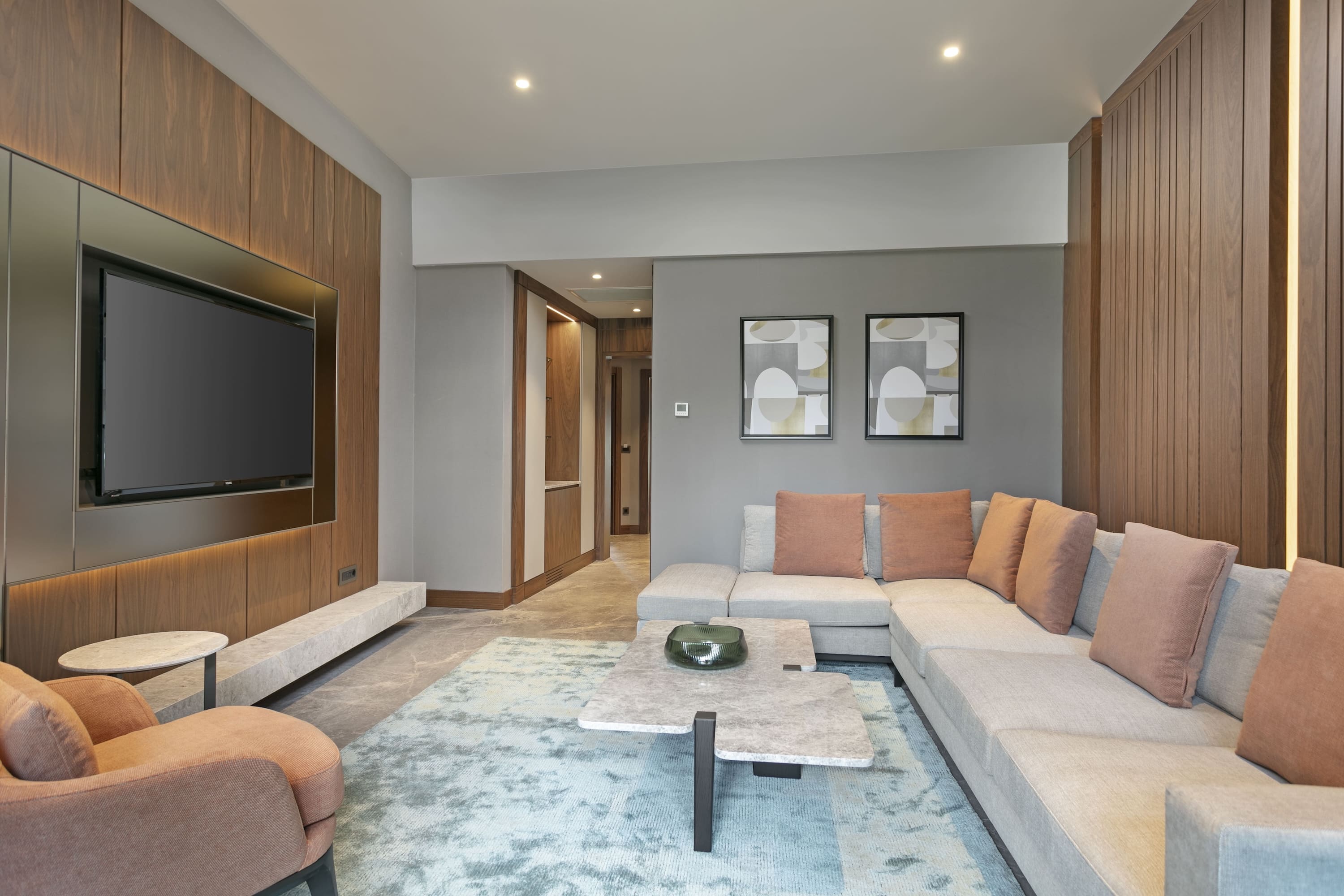Modern living room in the Rixos Sungate Marine Suite, featuring a grey sectional sofa with terracotta pillows, a marble coffee table, and a large wall-mounted television set against stylish wood paneling