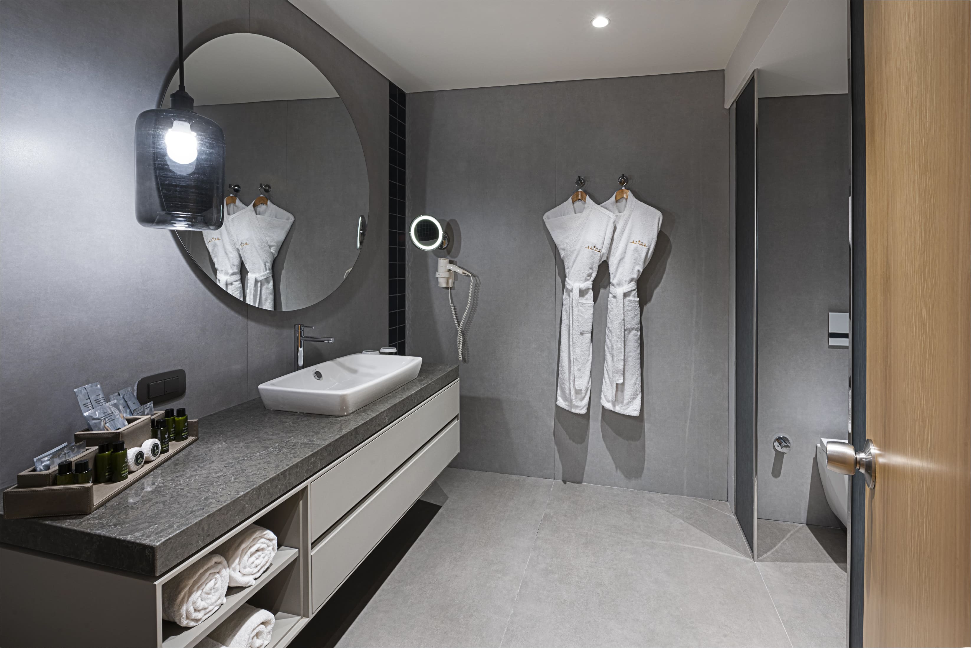 A modern bathroom in a Rixos Sungate Superior Room featuring a dark grey tiled wall, a large circular mirror, and a white vessel sink on a floating vanity. Two white bathrobes hang on the wall, and the countertop holds bathroom amenities and rolled towels are stored below.
