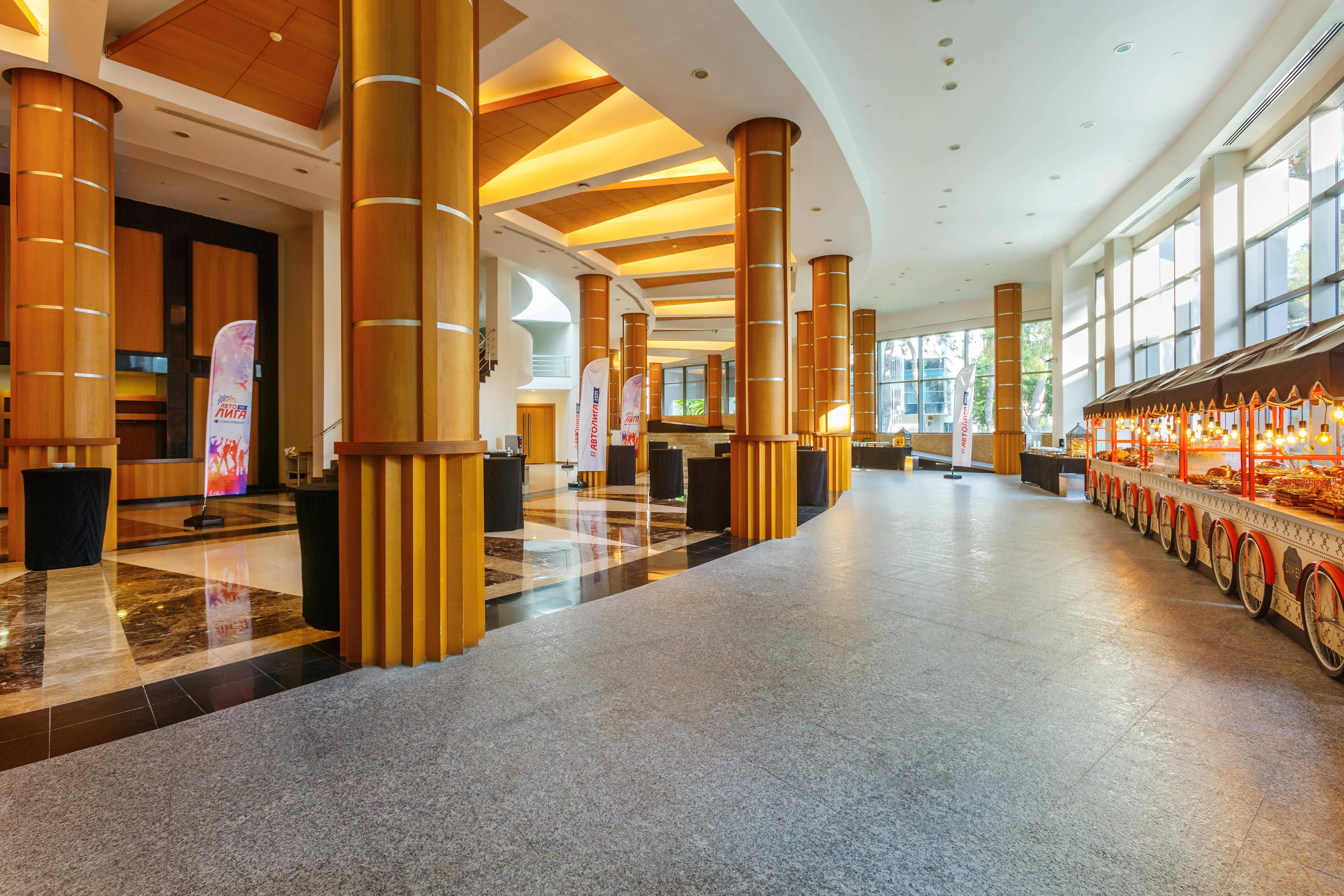 A wide shot of the brightly lit Spica Foyer at Rixos Sungate, a spacious area used for breaks and networking during a congress. The foyer features large wooden columns, a combination of dark marble and light grey tiled flooring, and high ceilings with recessed lighting. On the right, a row of tall windows lets in natural light. A decorative food service cart with a striped canopy is visible on the far right. Several tall, black draped cocktail tables and event flags are scattered throughout the Rixos space.