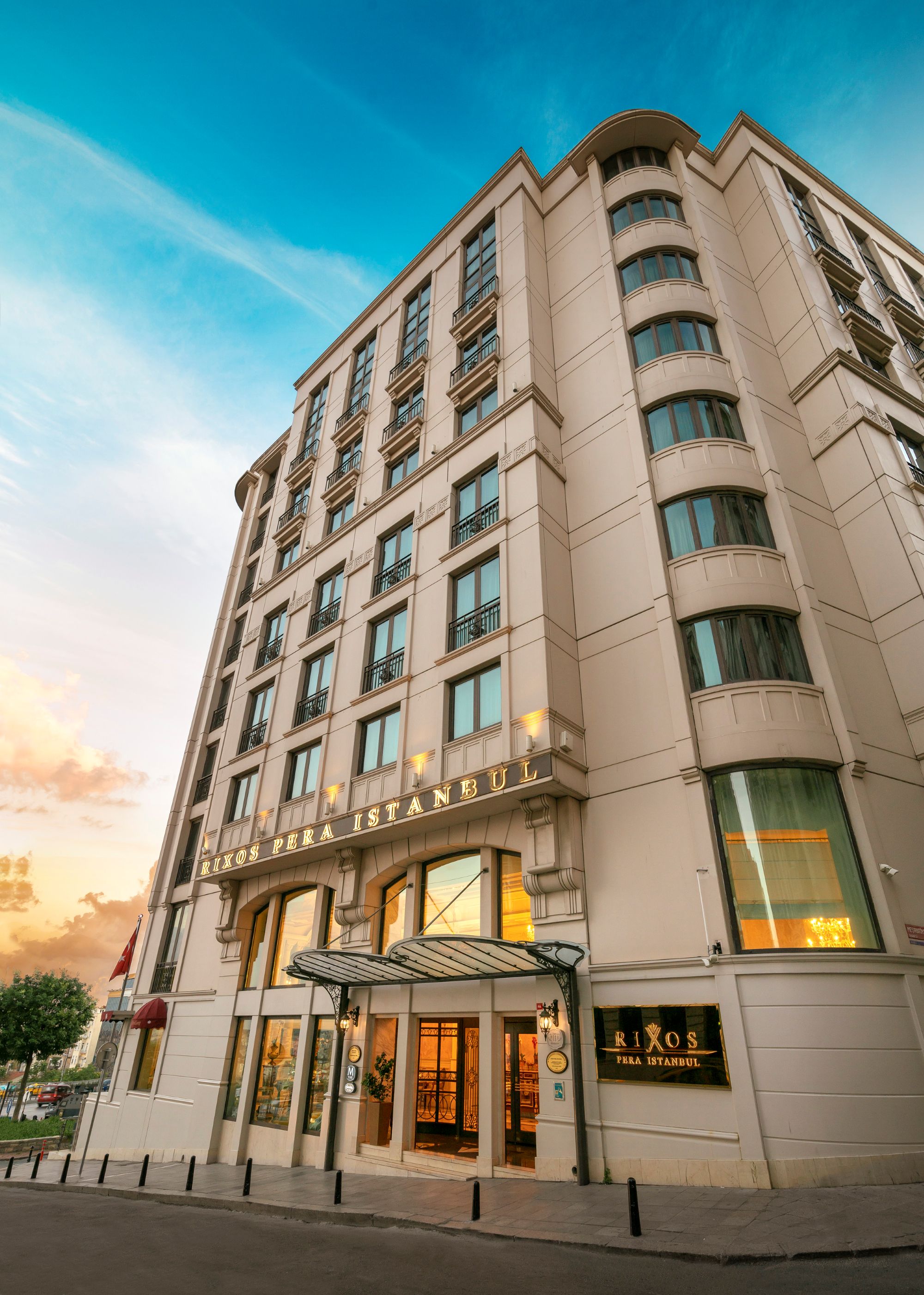 Rixos Pera İstanbul otelin dış cephesinin dramatik, yukarı doğru çekilmiş bir fotoğrafı. Yüksek, geleneksel taş bina, birkaç sıra pencereye ve alt kısmında kapalı bir ana girişe sahiptir. Bina, parlak mavi ve bulutlu bir gökyüzünün önünde yer almaktadır.